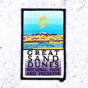 May include: Embroidered patch featuring a landscape scene with a blue sky, yellow sun, and purple and yellow mountains. The text "GREAT SAND DUNES NATIONAL PARK AND PRESERVE" is displayed in black and purple.