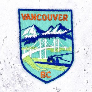 May include: A shield-shaped embroidered patch with a light blue background. It features the orange text VANCOUVER at the top and BC at the bottom. The design depicts a white suspension bridge, dark blue mountains, and green landscape elements.