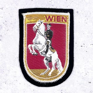 Op de afbeelding: Geborduurde patch met een wit paard dat steigerend staat, bereden door een persoon in uniform. De achtergrond is bordeauxrood met het woord "WIEN" in goud. De patch heeft een gouden rand en een zwarte rand.