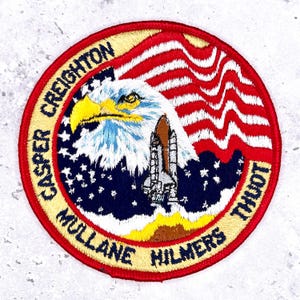 May include: Circular embroidered patch with an eagle, space shuttle, and the American flag design. The patch has a red border with text: "CASPER CREIGHTON MULLANE HILMERS THUOT". The eagle features yellow and blue detailing.