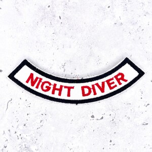 May include: A curved, embroidered patch featuring a white background with a thick black border. The text NIGHT DIVER is stitched in bold, red, capital letters across the centre of the patch, following the arc of the design.
