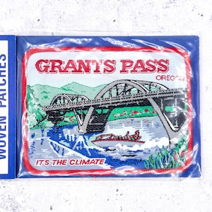 May include: A rectangular embroidered patch with a red border featuring a bridge over a river, a boat, and green hills. The text reads GRANT'S PASS OREGON and IT'S THE CLIMATE. The patch is enclosed in a clear plastic sleeve with a blue backing.
