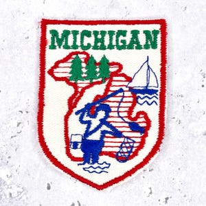 May include: A shield-shaped embroidered patch with a white background and a red border. The word MICHIGAN is stitched in green at the top. The design features a red outline of the state, green pine trees, a blue sailboat, and a blue fisherman.