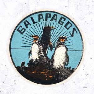 May include: A circular sticker featuring a light blue background with a sunburst design behind three penguins standing on dark rocks. The word GALAPAGOS is printed in bold black capital letters along the top edge of the circular border.