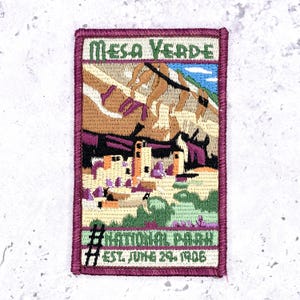 May include: A rectangular embroidered patch featuring a landscape of Mesa Verde National Park. The design includes cliff dwellings, rock formations, and a blue sky. Text reads MESA VERDE, NATIONAL PARK, and EST. JUNE 29, 1906. The border is dark purple.