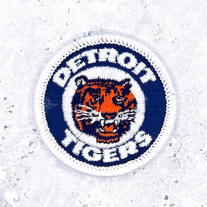 Puede incluir: Un parche bordado circular que presenta una cabeza de tigre en naranja y negro con bigotes blancos. El diseño está sobre un fondo azul oscuro con el texto blanco DETROIT TIGERS arqueado alrededor del centro, bordeado por costuras blancas.