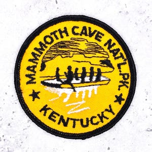 Op de afbeelding: Een ronde, gele patch met zwarte rand met de tekst "MAMMOTH CAVE NAT'L PK. KENTUCKY". De patch heeft een illustratie van mensen in een boot, met een grotomtrek erboven.