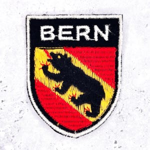 May include: A shield-shaped embroidered patch featuring the word BERN in white capital letters on a black background. Below the text, a diagonal yellow stripe crosses a red field, with a black bear silhouette centered on the shield design.