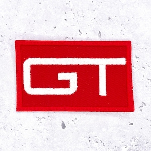 Op de afbeelding: Een rechthoekige rode patch met een wit geborduurd "GT" ontwerp. De patch heeft een rode rand en is waarschijnlijk bedoeld voor toepassing op kleding of accessoires. Het ontwerp is eenvoudig en gedurfd.