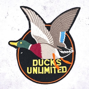 May include: Embroidered patch featuring a flying duck with outstretched wings. The duck has a green head, red chest, and gray and brown wings. The patch is in a black circle with the words "DUCKS UNLIMITED" in yellow.