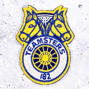 May include: An embroidered patch featuring two yellow horse heads facing outward above a circular emblem. The emblem contains a dark blue wagon wheel, the text TEAMSTERS in blue, and the number 182 in blue on a white background with yellow trim.