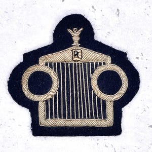 May include: A navy blue embroidered patch depicting a Rolls-Royce car emblem. The patch has a black felt background with gold thread detailing the car's grill, headlights, and hood ornament.