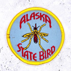 May include: A circular embroidered patch with a light blue background and a bright yellow border. The patch features a yellow and red mosquito graphic in the center with the red text ALASKA arched above and the red text STATE BIRD arched below.