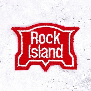 May include: A red embroidered patch featuring the text "Rock Island" in white. The patch has a distinctive shape with a white border. It is designed to be sewn onto garments or accessories.