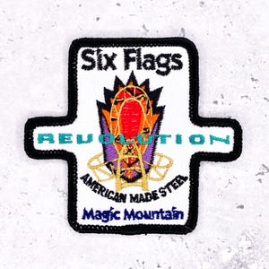 May include: Embroidered patch featuring the Six Flags Magic Mountain logo. The patch has a white background with the words "Six Flags," "Revolution," "American Made Steel," and "Magic Mountain." The design includes a colorful roller coaster image, outlined in black.