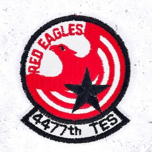 May include: Embroidered patch with a red eagle inside a red circle, with "RED EAGLES" above. A black star is below the eagle. The text "4477th TES" is in a black banner below the circle. The patch is circular.