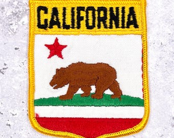May include: Embroidered patch with the California state flag design. The patch has a yellow border and the word "CALIFORNIA" in black. The design includes a brown bear, a red star, and the state colours of red, white, and green.