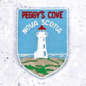 Op de afbeelding: Geborduurde patch met een vuurtoren met een rood dak, tegen een lichtblauwe lucht. De tekst "PEGGY'S COVE NOVA SCOTIA" is boven en onder de vuurtoren afbeelding gebogen. De patch heeft een schildvorm met een witte rand.