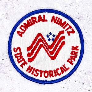 May include: A circular patch with a white background, a blue border, and red and blue text. The text reads "ADMIRAL NIMITZ STATE HISTORICAL PARK." A stylized red and white "N" shape is in the center, with a blue star design above it.