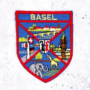 May include: Embroidered patch with a blue background and a red border. The patch features the word "BASEL" in yellow, along with illustrations of a bridge, buildings, and animals. A Swiss flag and a German flag are also depicted.