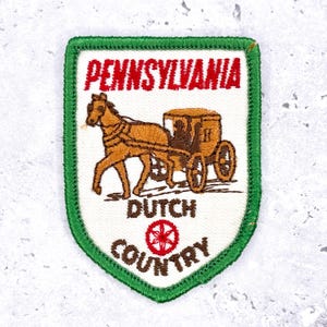 May include: Embroidered patch with a green border and a white interior. The patch features the word "PENNSYLVANIA" in red, above a brown horse-drawn carriage. Below the carriage, the words "DUTCH COUNTRY" are displayed in brown.