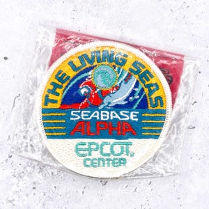 May include: A circular embroidered patch featuring a blue, yellow, and white design with the text THE LIVING SEAS SEABASE ALPHA EPCOT CENTER. The graphic depicts a stylized dolphin and a circular wave icon. The patch is sealed in a clear plastic bag.