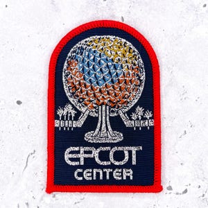 Peut inclure: Un écusson brodé rectangulaire avec un bord supérieur arrondi et une bordure rouge vif. Le fond bleu foncé présente un graphique de sphère géodésique blanc, bleu et orange au-dessus du texte blanc qui indique EPCOT CENTER en majuscules.