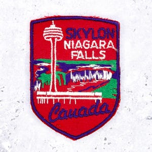 Op de afbeelding: Een schildvormige geborduurde patch met een rode achtergrond met de Skylon Tower, Niagara Falls en de tekst Skylon Niagara Falls Canada. Het ontwerp bevat groene, witte en paarse draaddetails die het landschap weergeven.