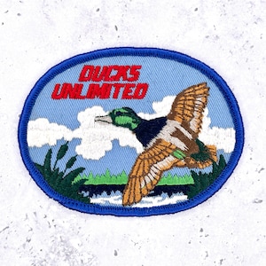 Pode incluir: Patch bordado oval com borda azul. O design apresenta um pato voador contra um céu azul claro com nuvens brancas. O texto "DUCKS UNLIMITED" é vermelho. Inclui juncos verdes e uma linha d'água.