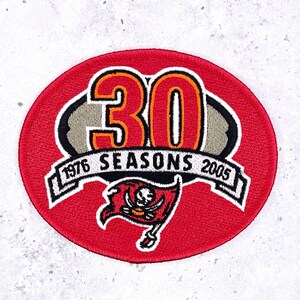 Puede incluir: Un parche bordado rojo circular con el número "30" en naranja y blanco, y las palabras "1976 SEASONS 2005". El parche también presenta una bandera pirata con una calavera y huesos cruzados.