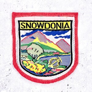 Pode incluir: Patch bordado com borda vermelha e design de paisagem. O patch apresenta a palavra "SNOWDONIA" acima de uma cena de montanha com um lago, árvore e um primeiro plano amarelo e verde. O patch é retangular com cantos arredondados.