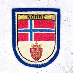 Peut inclure: Écusson rectangulaire avec une bordure bleue et un motif blanc, rouge et bleu. La partie supérieure porte le mot "NORGE" en lettres dorées. En dessous, un drapeau rouge et bleu et un écusson rouge avec un lion doré.
