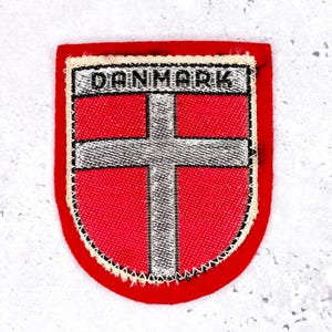 May include: A red and white embroidered patch with the word "DANMARK" at the top. The patch features a white cross on a red background, with a red border and a decorative stitch.