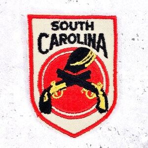 May include: Embroidered patch featuring the words "SOUTH CAROLINA" in black, above a red circle with crossed black pistols and a yellow and black design. The patch has a cream-colored border and a red outline.