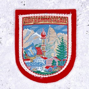 May include: A red and white embroidered patch with the text "OBERAMMERGAU". The patch features a detailed landscape scene with mountains, a church, and houses. The patch has a red border.