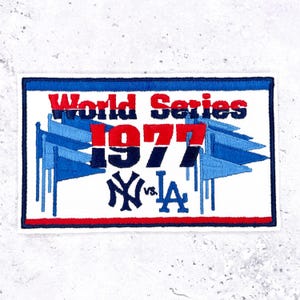 May include: Embroidered patch with a white background, blue border, and the text "World Series 1977." The patch features blue pennants and the logos of the New York Yankees and the Los Angeles Dodgers.