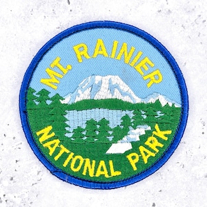 Pode incluir: Remendo bordado circular com o Parque Nacional Mount Rainier. O remendo tem uma borda azul e um fundo azul claro. O design inclui uma montanha, um lago e árvores, com o texto "MT. RAINIER NATIONAL PARK" em amarelo.