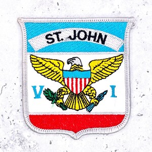 May include: A shield-shaped embroidered patch featuring a light blue top section with the text ST. JOHN in black. The center displays a yellow eagle with a shield on its chest, holding a green olive branch and blue arrows. The bottom is red.