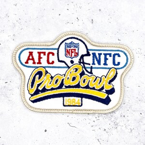 May include: A vintage NFL Pro Bowl patch with a white background and a cream-colored border. The patch features the text "AFC" and "NFC" in red and blue, respectively, and a football helmet with the NFL logo. The words "Pro Bowl" are in yellow script, with the year "1984" below.
