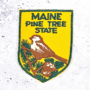 May include: A shield-shaped embroidered patch with a yellow background and a green border. It features a brown and white bird perched on a green pine branch with a brown pinecone. The text MAINE PINE TREE STATE is embroidered in green.