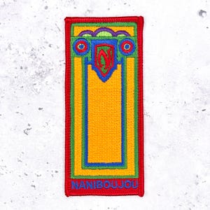 May include: A rectangular embroidered patch featuring a vibrant yellow centre bordered by green and blue lines. The top section displays a red letter N inside a blue shield, flanked by blue circles. The word NANIBOUJOU is stitched in blue at the bottom.
