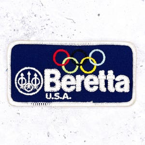 May include: A rectangular embroidered patch with a dark blue background and a white stitched border. It features the white text Beretta U.S.A., a circular logo with three arrows, and five interlocking rings in red, black, blue, yellow, and green.