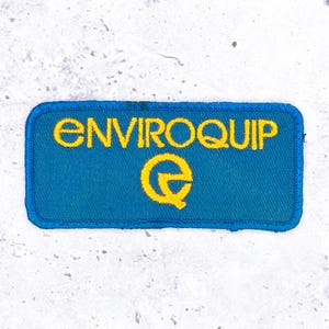 May include: A rectangular embroidered patch featuring a blue background with a dark blue stitched border. The text ENVIROQUIP is embroidered in yellow capital letters across the top, with a stylized yellow letter Q logo positioned below the text.