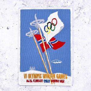 May include: A rectangular embroidered patch with a light blue background featuring two crossed ski poles, a white flag with the five Olympic rings, and a Norwegian flag. The text reads VI OLYMPIC WINTER GAMES 14-26 FEBRUARY OSLO NORWAY 1952.