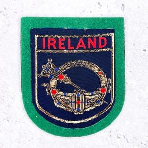 May include: A green-edged patch with the word "IRELAND" in red above a detailed gold and blue emblem. The emblem is set against a dark blue background, with intricate gold detailing and red accents.