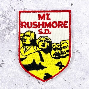 May include: Embroidered patch depicting Mount Rushmore National Memorial. The patch has a white background with a red border. The text "Mt. Rushmore S.D." is at the top. The image shows the faces of the four U.S. presidents.