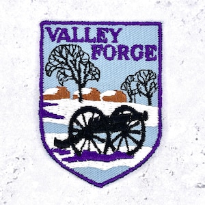 May include: A shield-shaped embroidered patch with a light blue background and a purple border. It features the text VALLEY FORGE in purple, black silhouettes of two trees, brown log structures, and a black cannon on a white snowy ground.
