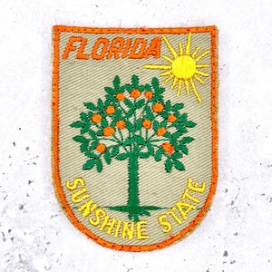 Puede incluir: Un parche bordado en forma de escudo con fondo color canela claro y borde naranja. Presenta un naranjo con hojas verdes y frutos naranjas, un sol amarillo y el texto FLORIDA en la parte superior y SUNSHINE STATE en la parte inferior.
