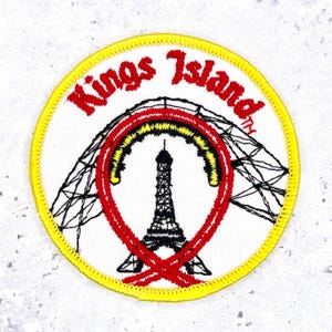 May include: A circular embroidered patch with a yellow border on a white background. It features the red text Kings Island above a black graphic of the Eiffel Tower and a roller coaster track loop outlined in red and yellow thread.