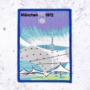 May include: A rectangular embroidered patch featuring a purple sky, a white Olympic tower, and stylized tent structures. The text reads München 1972 26.8.-10.9. with the Olympic rings logo. The patch has a thick blue border and measures in a vertical orientation.